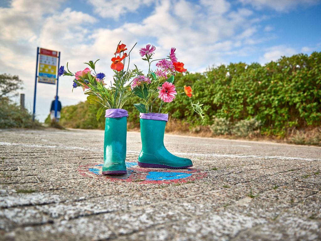 Blaue Gummistiefel mit Blumen bepflanzt stehen auf einem Weg zum Strand
