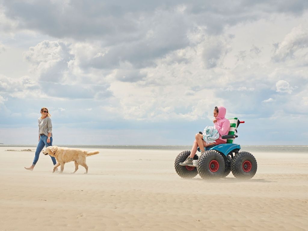 Das Foto zeigt eine junge Frau mit einem golden Retriever, die mit einer jungen Frau im Strandmobil am windigen Strand von Langeoog spazieren geht